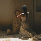Woman sitting on the floor weaving a basket in a dimly lit room.