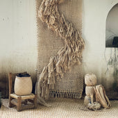 Textile art piece made of natural fibers on a wall with a woven rug and small stool in the foreground.