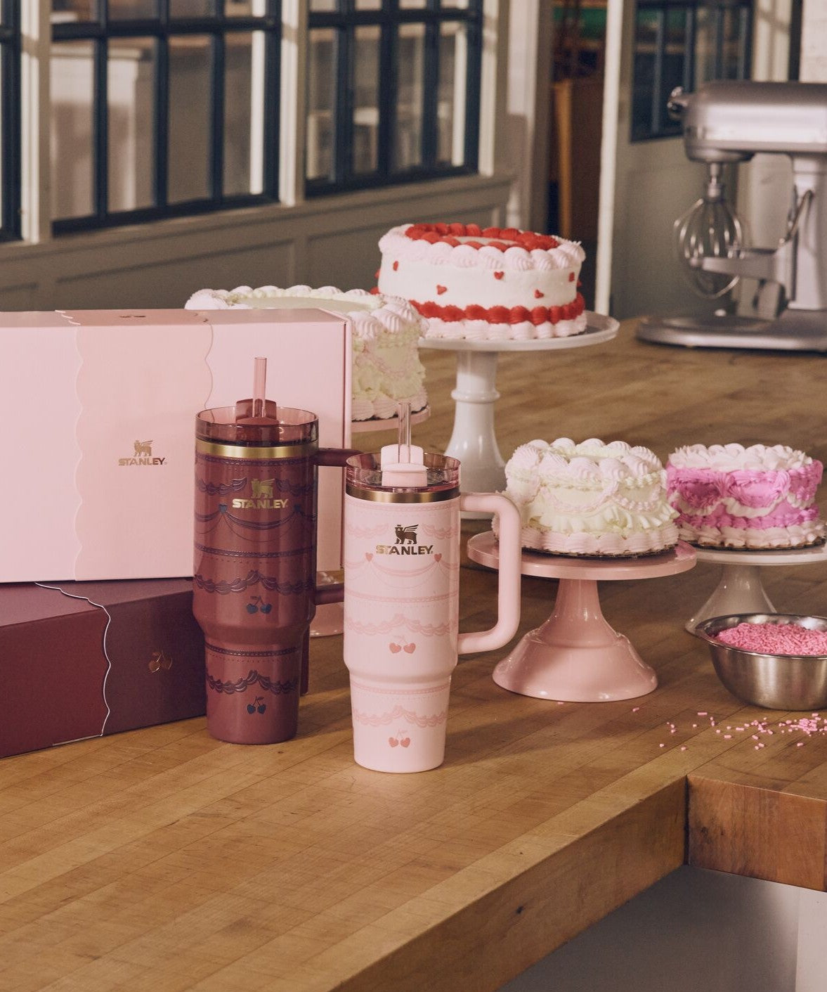 Two pink tumblers on a wooden surface with cakes in the background