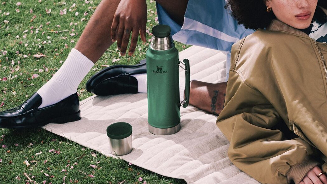 Two people sitting on a blanket in a park with a green thermos and phone visible.