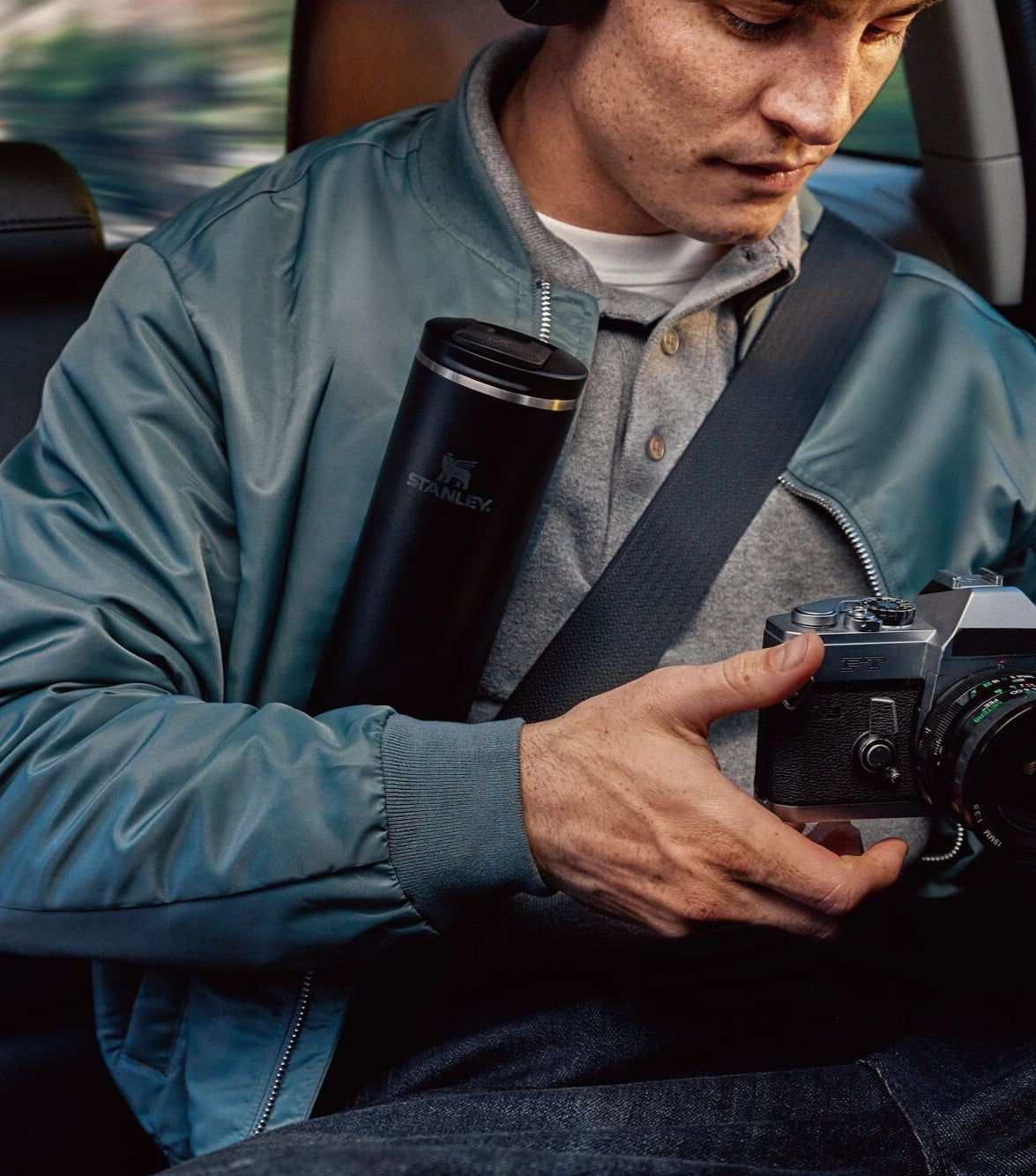 Person sitting in a car holding a camera and a thermos, wearing a blue jacket.